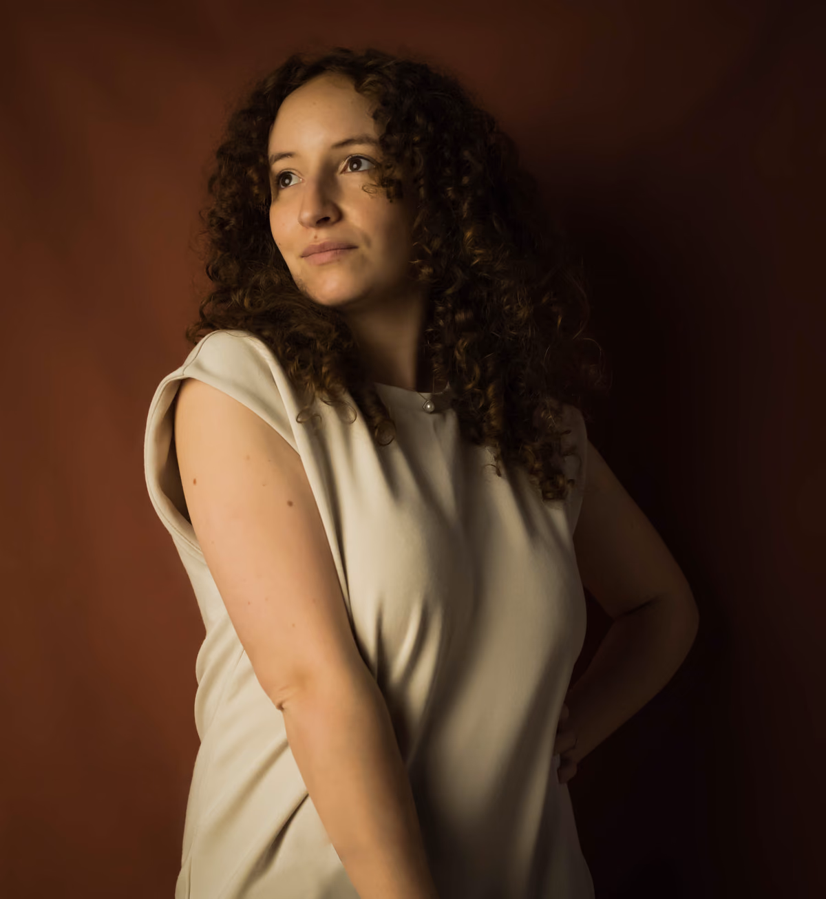 Sarah Fenehari, de trois quarts, en robe claire sur un fond brun chaud
